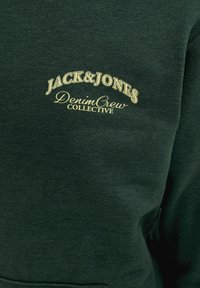 Tissu vert foncé avec texte crème indiquant "JACK & JONES Denim Crew Collective" sur la zone de la poitrine d'un vêtement.