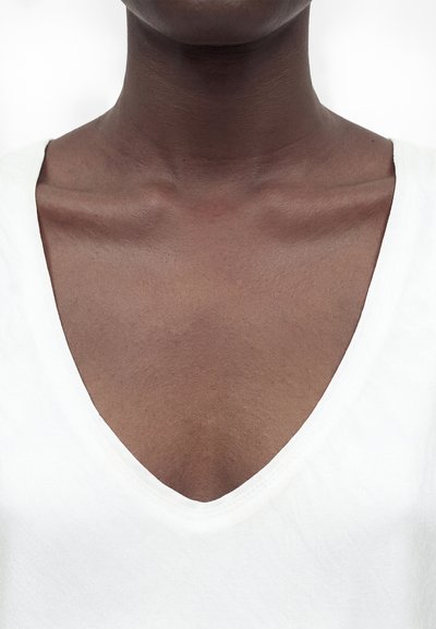 Κοντινό πλάνο του λαιμού και του άνω μέρους του στήθους ενός ατόμου με σκουρόχρωμο δέρμα που φοράει λευκό μπλουζάκι με V-λαιμόκοψη σε απλό φόντο.