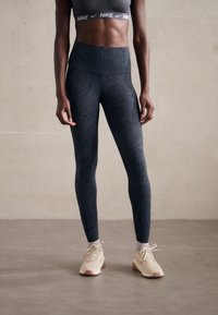 Schwarze Leggings mit einem strukturierten Wirbelmuster, hohem Bund, kombiniert mit einem grauen Sport-BH und beigen Sneakers mit pinken Akzenten.