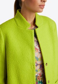 Bright green textured coat with a high collar and front fastening. It features a soft, fluffy fabric and gold-tone buttons, layered over a floral print dress.