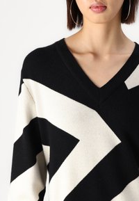 Pull à motif noir et blanc avec un col en V, présentant un design en zigzag audacieux et une texture douce. Une vue rapprochée révèle les détails du tricot fin.