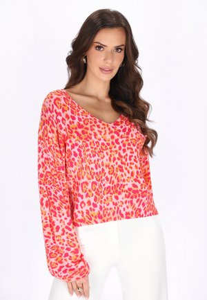 Kobieta z długimi brązowymi falistymi włosami, nosząca złote kolczyki koła, różową bluzkę w panterkę z długim rękawem oraz białe spodnie, pozująca na białym tle.