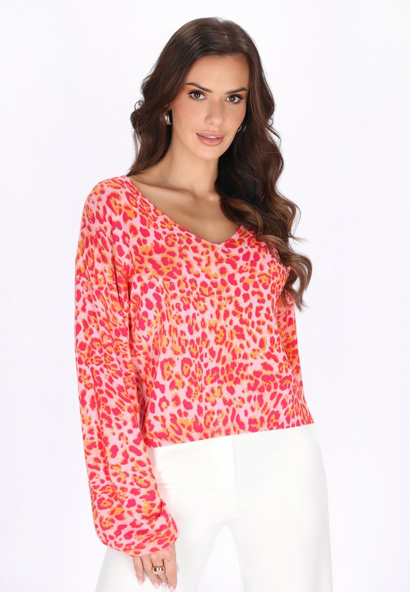 Frau mit langem, braunem, gewelltem Haar trägt goldene Creolen, ein pinkes Leopardenmuster-Langarmshirt und weiße Hose, posiert vor weißem Hintergrund.