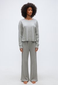 Γυναίκα με σγουρά μαλλιά, που φοράει χαλαρό γκρι μπλουζάκι με μακριά μανίκια και γκρι παντελόνι φαρδύ, στεκόμενη ξυπόλητη μπροστά σε ένα απλό φόντο.