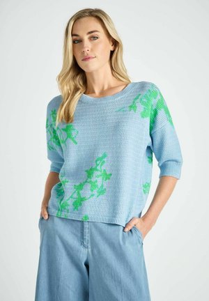 Blonde vrouw die een lichtblauwe gebreide trui met groene bloemmotieven en losse lichtblauwe broek draagt, staand met handen in de zakken.