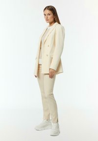 Beiger Blazer mit Nadelstreifen und doppelter Knopfleiste, kombiniert mit einem beigen Oberteil und passenden schmal geschnittenen Hosen. Weiße Sneakers runden das Outfit ab.