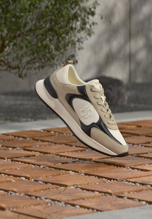 Beige en zwarte sneaker met veters, zwevend boven houten tegels buiten met een wazige achtergrond van groen en beton.