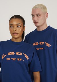 Blå bomulds t-shirts med fedt, orange grafisk tekst og stjernetema. Løs pasform, rund hals, korte ærmer, ensfarvet baggrund.