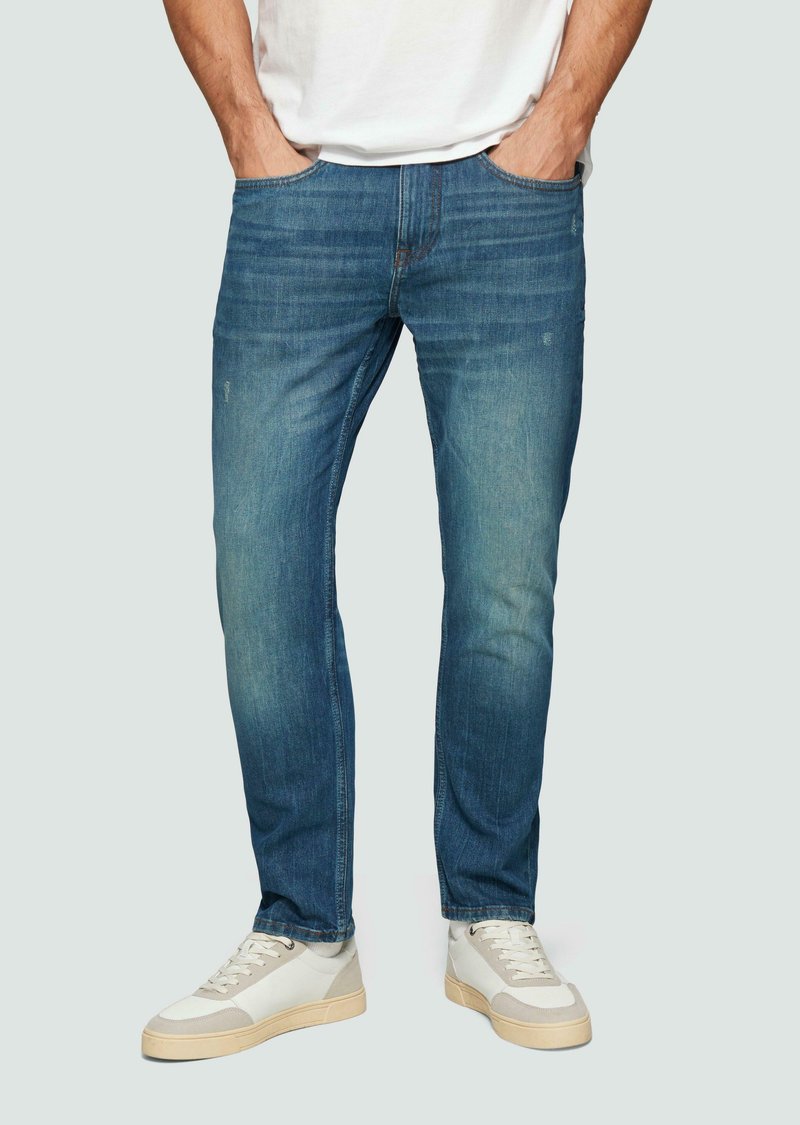 Man in een blauwe slim-fit jeans en witte sneakers, met handen in zijn zakken, staande tegen een effen lichte achtergrond.