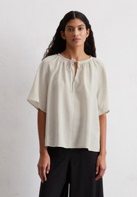 Helle beige Bluse mit kurzen Ärmeln, rundem Ausschnitt und einer Herzformöffnung, die mit einem Band gebunden ist. Textil mit sanften Wellenstrukturen.