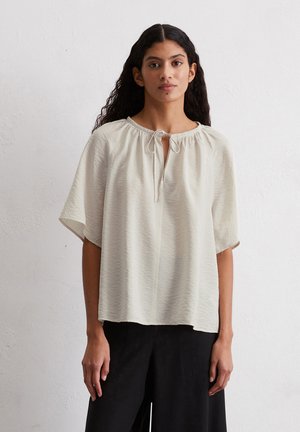 Light beige blouse with short sleeves, a round neckline, and a keyhole opening tied with a string. Textured fabric with soft waves.