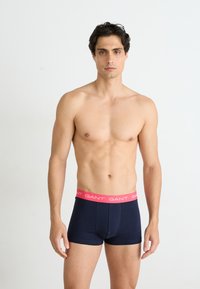 Marineblauwe boxershorts met een roze elastische tailleband waarop de merknaam "GANT" prominent in witte letters is weergegeven.