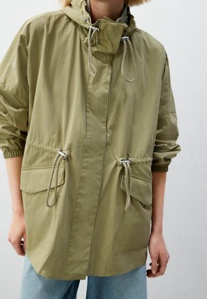 Personne portant une veste à capuche vert olive avec des cordons au niveau du cou et de la taille, et de grandes poches avant à rabat, associée à un jean bleu clair.