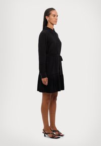 Melna kleita ar pogu priekšpusi, plisētu svārku un jostu viduklī. Nēsāta ar melniem siksnu papēžiem. Gluda tekstūra, precīzs piegriezums.