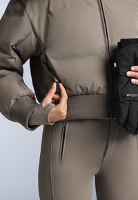 Matt barna színű pufi dzseki bordázott mandzsettákkal, rövidített fazonnal és cipzáros zsebekkel. Fekete kesztyűk kézben tartva.