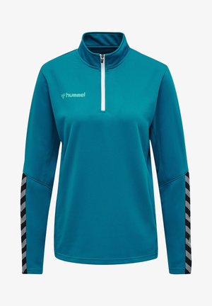 Teal langærmet træningstrøje med halvdels-zipkrave, tekstureret stof og sort-hvide chevronmønstrede detaljer på ærmerne.