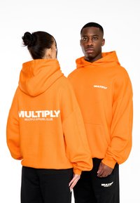 Sudaderas naranjas con un corte relajado, que presentan el texto "MULTIPLY" en blanco en el pecho y la espalda. Se llevan pantalones negros con las sudaderas.