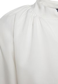 Vêtement blanc avec un détail de tissu froncé près du col montant et de la couture de l'épaule, montrant une texture lisse et des plis légers.