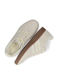 Baskets beige avec une tige en daim lisse, bandes latérales blanches, semelle en caoutchouc marron et détails de logo embossés sur le côté extérieur.