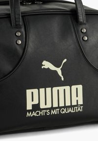 Sort lædertaske med cremefarvet Puma-logo og tekst "PUMA MACHT'S MIT QUALITÄT," med to lodrette håndtag med metalnitter.