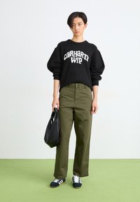 Czarny sweter dzianinowy z białym logo "Carhartt WIP", oliwkowe spodnie, czarna torba i niebieskie trampki z białymi paskami na zielonej podłodze.