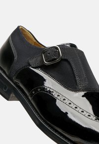 Schwarze Lederschuhe mit glänzender Oberfläche, ausgestattet mit einem Obermaterial aus Wildleder, dekorativen Perforationen und einem metallischen Schnallenakzent.