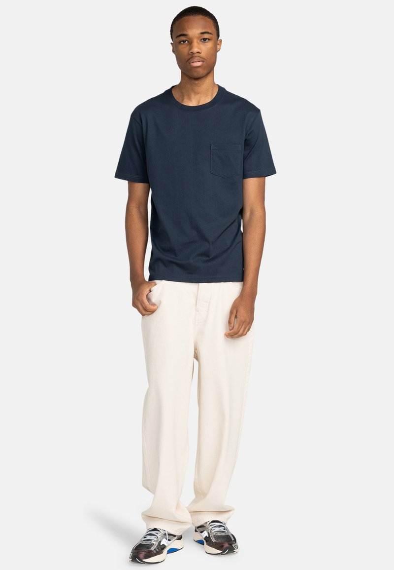 Navy katoenen t-shirt met een ronde hals en een borstzak, gecombineerd met losse crèmekleurige broeken en geblokte sportschoenen.