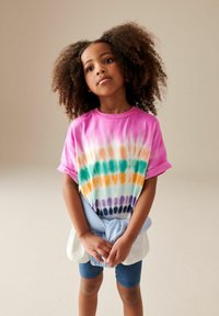 Koszulka z bawełny w technice tie-dye w różowych, pomarańczowych, zielonych i fioletowych wzorach. Rolowane rękawy, noszona z niebieskimi szortami i jasnoniebieskim związanym swetrem.