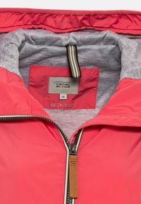 Veste rouge recyclée avec une doublure grise, dotée d'une fermeture éclair à l'avant et d'une étiquette de suspension en tissu. L'étiquette indique "camel active" et taille 36.