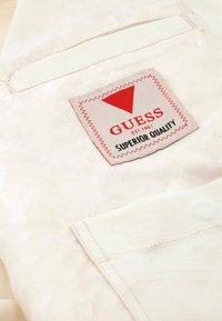 Cream-colored fabric with a textured pattern. Features a red triangular logo patch with "GUESS" and "SUPERIOR QUALITY" text.
