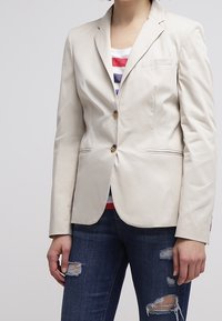 Femme portant un blazer beige boutonné sur un haut rayé rouge, blanc et bleu, associé à un jean foncé déchiré, sur un fond uni.