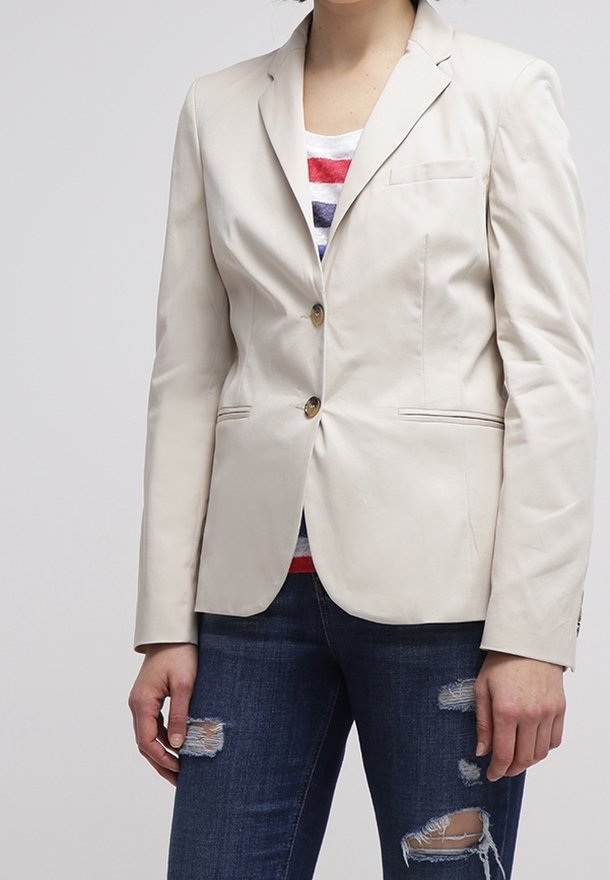 Femme portant un blazer beige boutonné sur un haut rayé rouge, blanc et bleu, associé à un jean foncé déchiré, sur un fond uni.