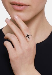 Srebrny pierścionek z czarnym wzorem krzyża, ozdobiony przezroczystymi kamieniami, zaprezentowany na dłoni dotykającej podbródka o neutralnym odcieniu skóry.