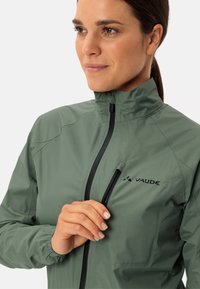 Lichte groene ritsjack met staande kraag, textuurstof en een zwarte ritszak. Heeft een klein zwart logo op de borst.