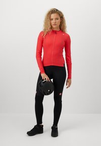 Femme en veste de cyclisme rouge et leggings noirs tenant un casque de vélo noir, se tenant contre un fond clair.
