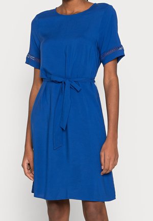 Femme portant une robe bleu roi longueur genoux avec manches courtes, ceinture nouée à la taille et bordure en dentelle aux extrémités des manches.