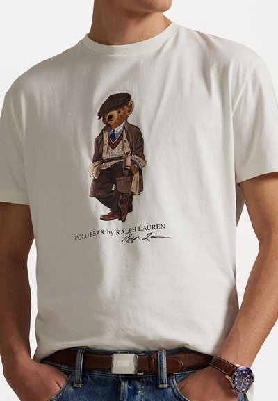 T-shirt en coton blanc avec une illustration détaillée d'un ours en costume, accompagné d'accessoires. Inclut un logo et un texte de marque.
