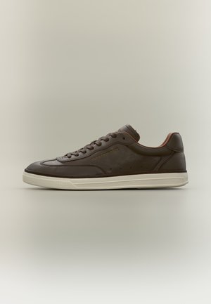 Niski, ciemnobrązowy skórzany sneaker z białą podeszwą i sznurówkami, widok z boku na beżowym tle.