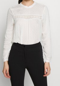 Blusa bianca a maniche lunghe in tessuto texturizzato, con un pannello in pizzo trasparente sul petto e scollatura rotonda, abbinata a pantaloni neri su misura.