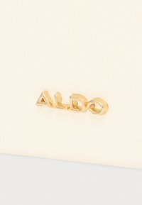 Le matériau de couleur crème présente un logo "ALDO" en relief doré. La surface a une texture subtile, mettant en valeur une finition lisse.