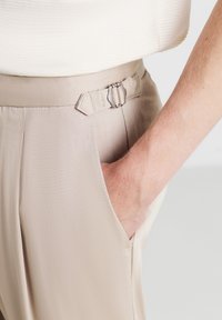 Beige maßgeschneiderte Hosen mit glatter Textur, mit einer seitlichen Tasche und einer doppelten Schnallenakzentuierung am Bund.