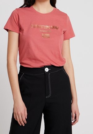 Frau trägt ein korallenfarbenes T-Shirt mit dem Text "EXPEDITION CAMP" und schwarze hoch taillierte Hosen mit weißen Knöpfen und Nahtdetails.