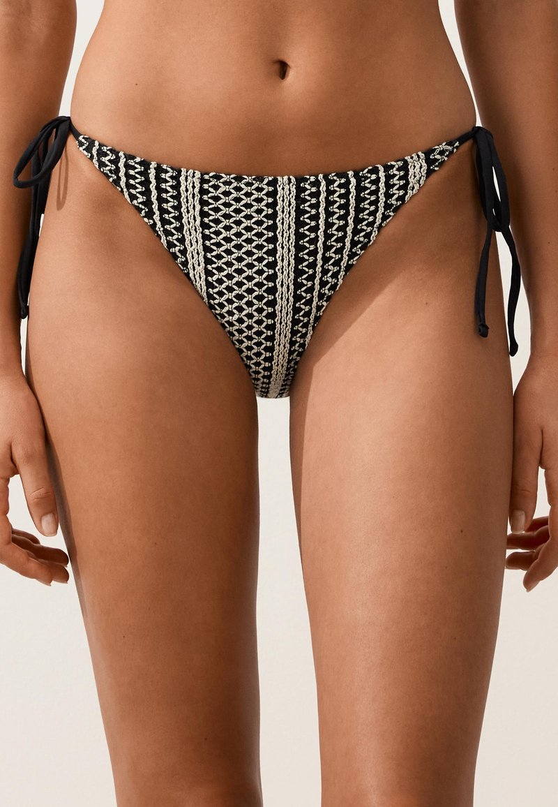 Femme portant un bas de bikini noir et blanc en crochet avec des liens sur les côtés, vue de la taille jusqu'à la mi-cuisse sur un fond neutre.