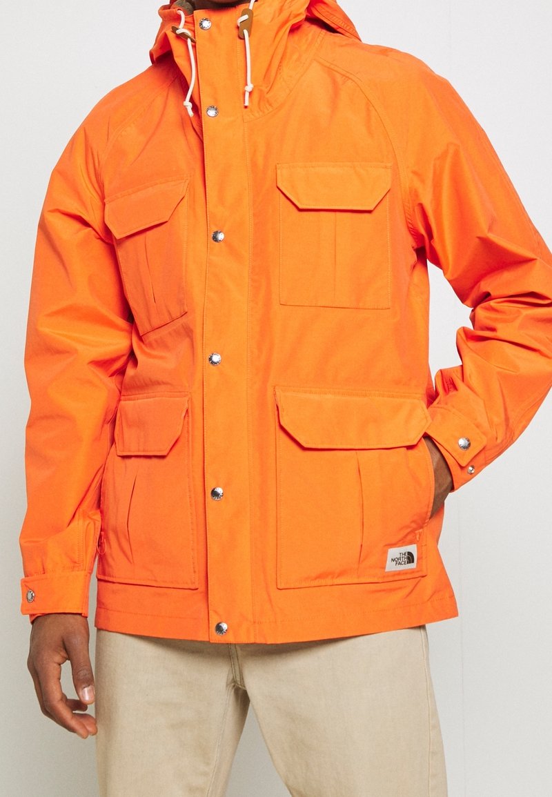 Jasně oranžová bunda vyrobená z hladkého, odolného materiálu. Má několik předních kapes, drukové zapínání a nastavitelnou kapuci s tkaničkami.