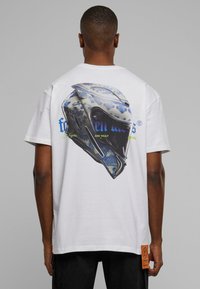 Weißes T-Shirt mit einem großen Grafikdesign eines blauen und weißen gemusterten Helms auf der Rückseite, akzentuiert mit mehrfarbigem Text und einem Textillabel.