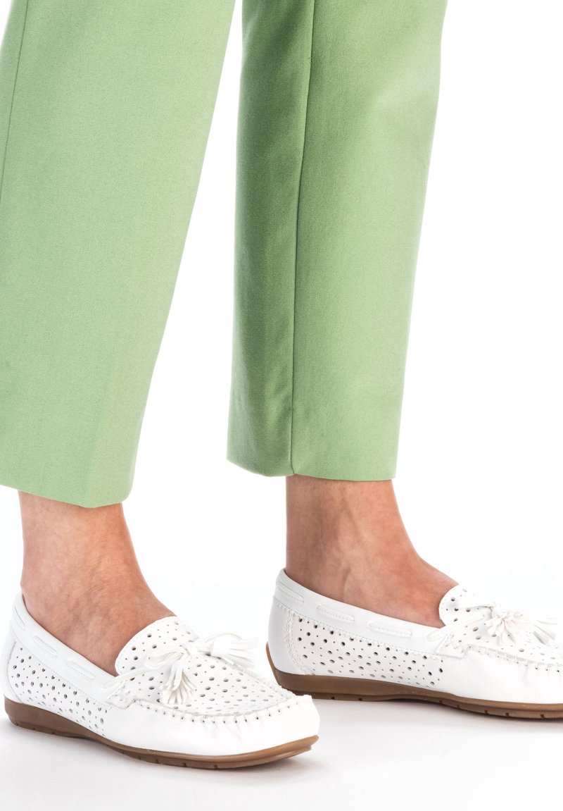 Chaussures blanches en cuir de style mocassin avec un design perforé, semelles plates et des glands décoratifs, associées à un pantalon court vert clair.