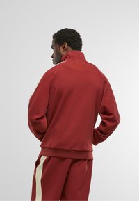 Muž v červenej teplákovej súprave s krémovými pruhmi stojí s rukami v vreckách, otočený chrbtom proti jednoduchému svetlému pozadiu.