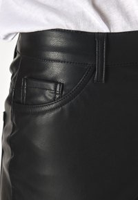 Svart fusk läderkjol med en slät textur, som har en böjd fickdetalj och synlig sömnad på midjebandet.