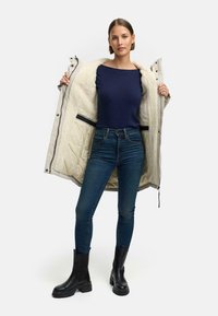 Beżowy ocieplany płaszcz z fakturowanym kremowym podszyciem, granatowa bluzka i ciemnoniebieskie dżinsy. Na nogach ma czarne botki na kostkę z masywną podeszwą.