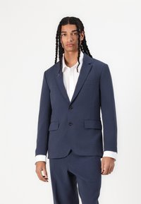 Marineblauwe colbert van gestructureerde stof, twee voorzakken, sluiting met één knoop, gecombineerd met een witte shirt met accentknopen.